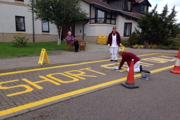 two men painting letters outside a care home, one looking to camera. An elderly woman watches from a nearby bench.
